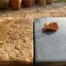 Natte getextureerde steentegel met waterdruppels naast een gladde geglazuurde tegel op een zonnig terras, met een herfstblad.
