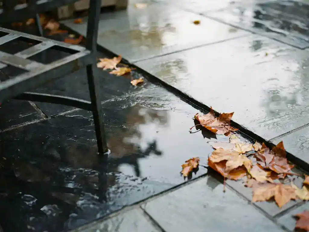Glanzende gepolijste stenen tegels op een natte buitenterras met regenwater, herfstbladeren en een tuinstoel op de achtergrond.