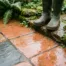 Natte terracotta en natuurstenen terrastegels met waterdruppels, modderige rubberlaarzen op de rand, omringd door weelderige tuinbeplanting.