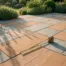 Terracotta en grijze buitentegels in geometrisch patroon op zonnig terras, met meetlint en wazige tuinbeplanting op de achtergrond.