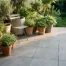 Grote keramische tegels met matte betonlook finish op een compact terras, met groene potplanten en een bistrostoel in warm middaglicht.