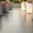 Glanzende betongrijs keramische vloertegels in strak gridpatroon op zonnig buitenterras met terrasstoelen op de achtergrond.